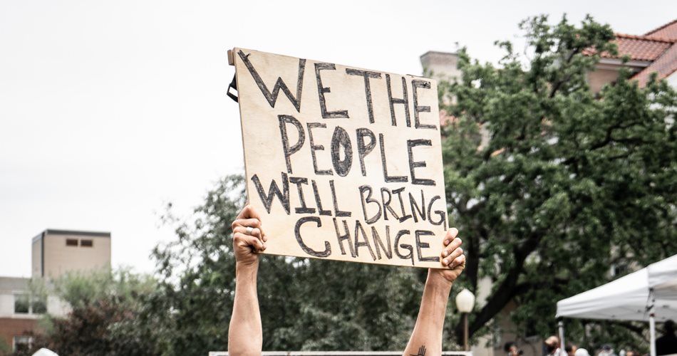 Young white man in the middle of a crowd holding a cardboard sign above his head that reads 'We the people will bring change'