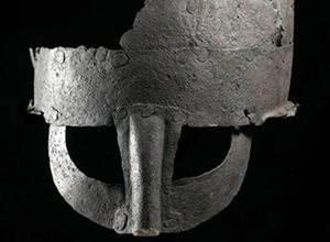 Photograph of a rusted and partially missing ancient helmet with a plain curved helm, noseguard and covers around the eyes against a black background.