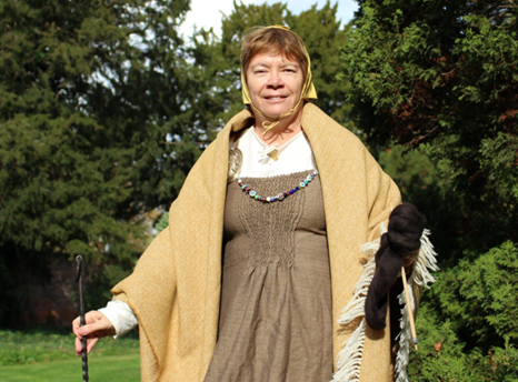 Photograph of a white woman with brown hair wearing traditional Viking dress standing outside in a wooded glade on a sunny day