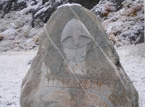 Photograph of a roughly triangular stone with a helmet and a row of runes curved on the flat face of the rock.