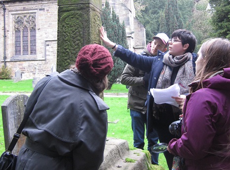 Four people standing by a grave outside a church looking in the direction of the middle figures up- and left-wards pointing finger (photo).