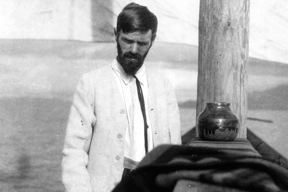 Black and white photographic portrait of DH Lawrence in a white suit jacket stood next to a wooden mast.