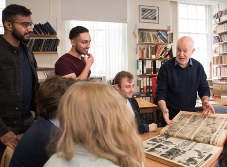 Diverse group of students clustered around a table in a library while an older white man shows them a large old book.