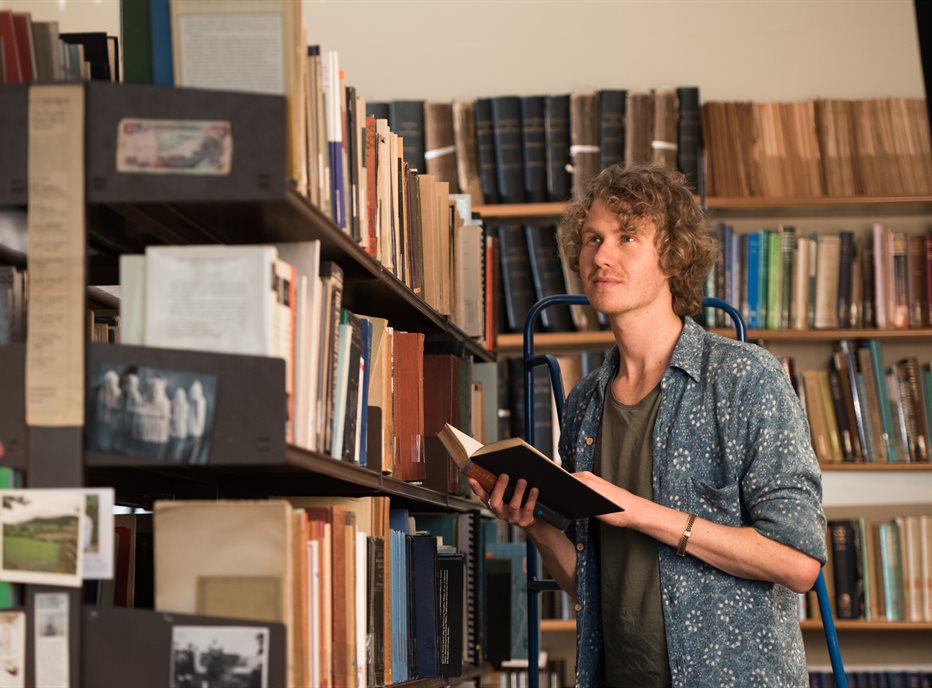 Photo of a young white male student holding a book in a room lined with bookshelves