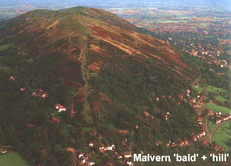 Aerial photograph of a large hill with brown grass on the top, bare trees on the lower slopes and red roofed houses scattered around its base.