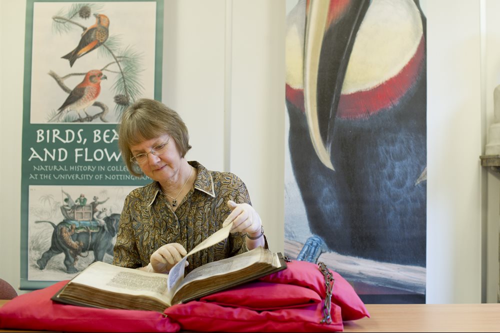 A white older lady sat in front of a banner about an event called 'Birds, Beasts and Flowers' carefully lifts a page in a very large old hardback book which is resting on a pile of red cushions.
