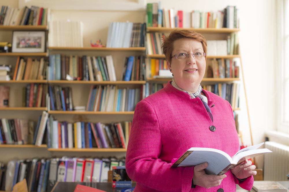 Dr Christina Lee wearing a bright pink jacket stood holding a book in her office, Trent Building, with shelves behind.