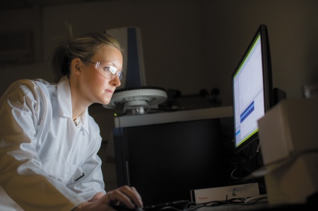 A researcher in white lab coat and goggles looks at a computer screen