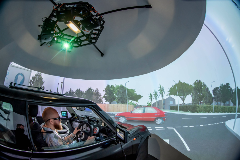 Research Assistant at the wheel inside the driving simulator showing them at a junction on the main screen with a blue car passing
