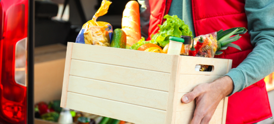 Courier holding crate with products near car outdoors, closeup. Food delivery service